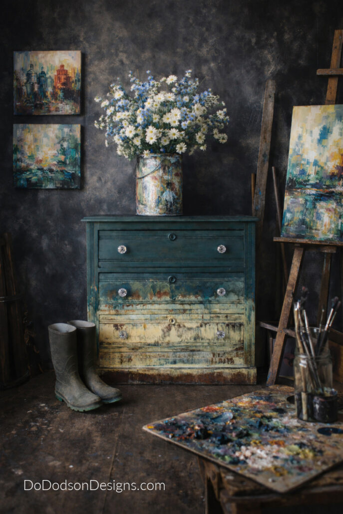 Hand painted artistic dresser with layered distressed paint finish styled in a moody art studio with dark walls and flowers in a paint can.