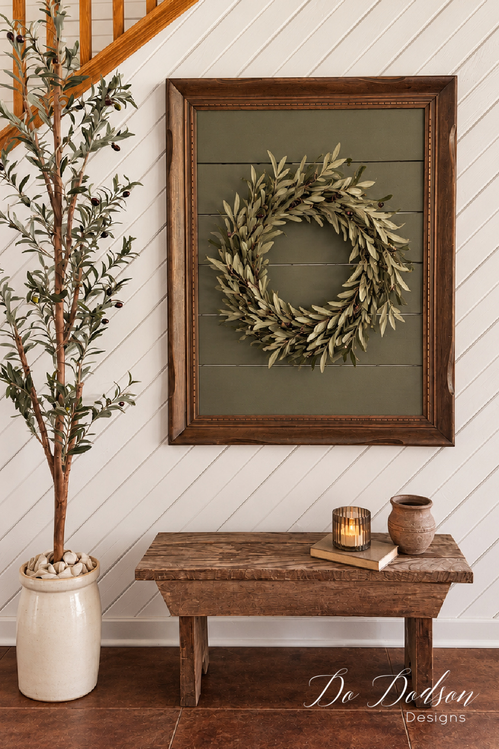 Dark wood picture frame with olive green faux shiplap and an olive leaf wreath, hanging above a rustic wood bench beside a potted olive tree.