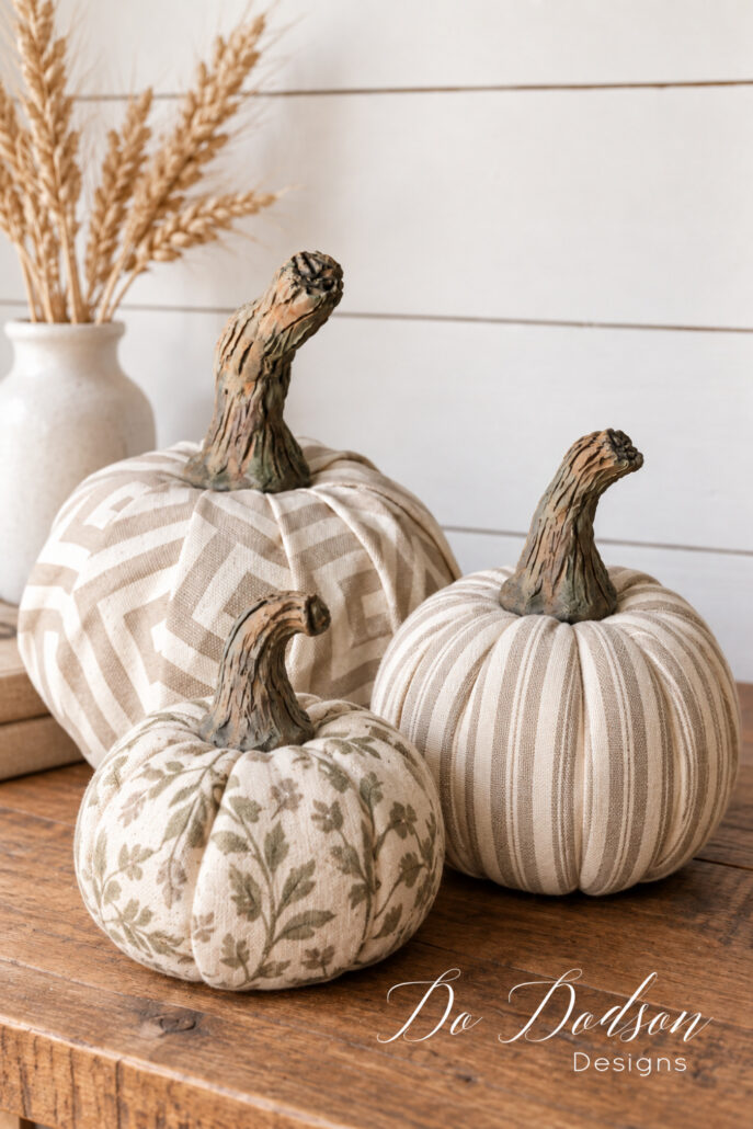 Three fabric pumpkins in geometric, striped, and floral patterns with realistic handmade clay stems on a farmhouse table