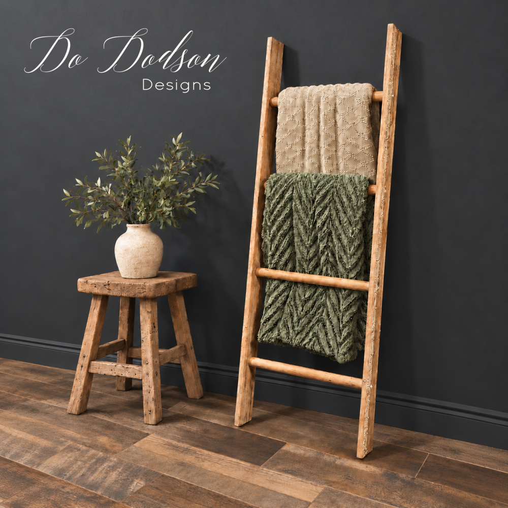 Rustic wooden blanket ladder displaying beige and olive green throws beside a wood stool with a vase of greenery against a dark charcoal wall.