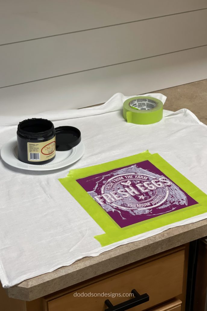 Remove the silk screen stencil backing (save for later) and place the sticky side down on the flour sack towel. I like to use painter's tape to add a border around the stencil to protect the fabric from runaway paint.