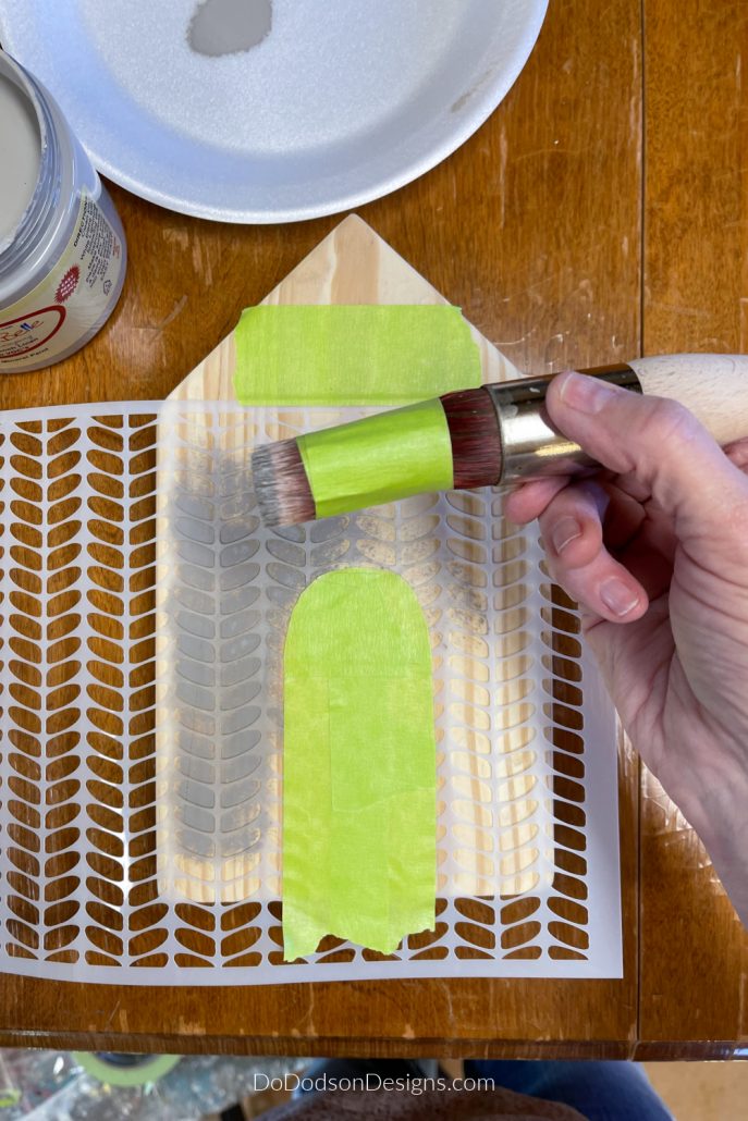 Lay the stencil over the wood block houses and use painter's tape to cover the areas where you want the outline of the doors or windows to be. Then secure the stencil in place using painter's tape to keep it from slipping as you stencil your design. 