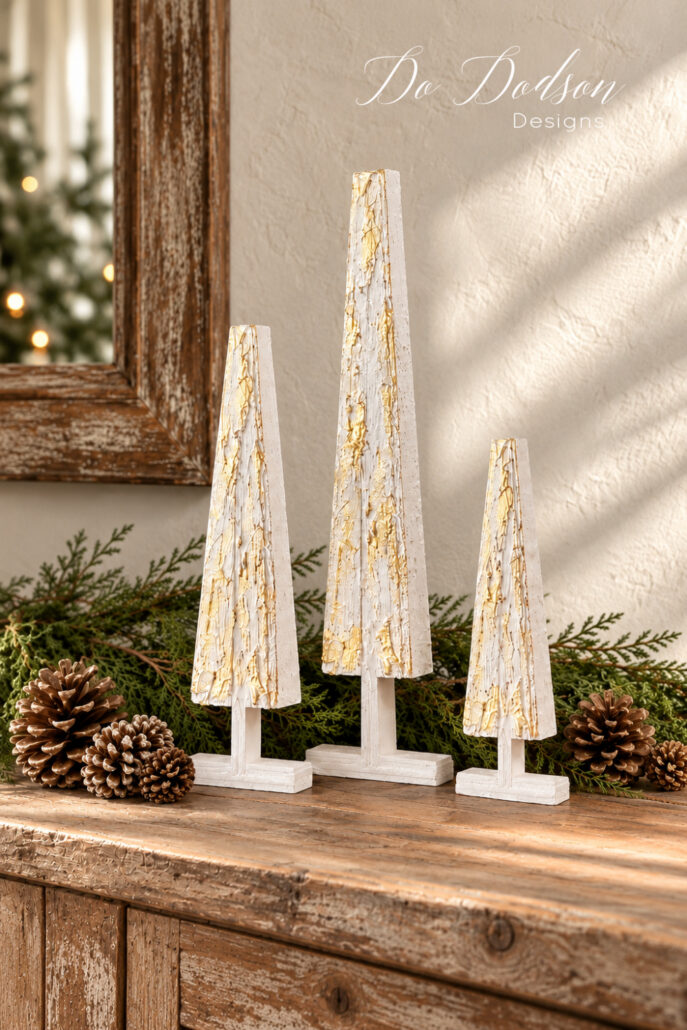 Three DIY wood shim Christmas trees of varying heights with textured white and gold finishes displayed on a rustic wooden table with evergreen branches and pinecones.