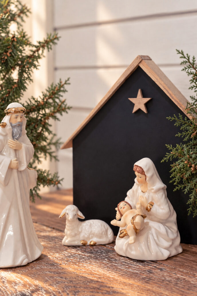 White ceramic Mary holding baby Jesus, Joseph, and a sheep displayed before a black DIY wooden nativity stable with a natural wood shim roof and matching star, surrounded by cedar greenery.