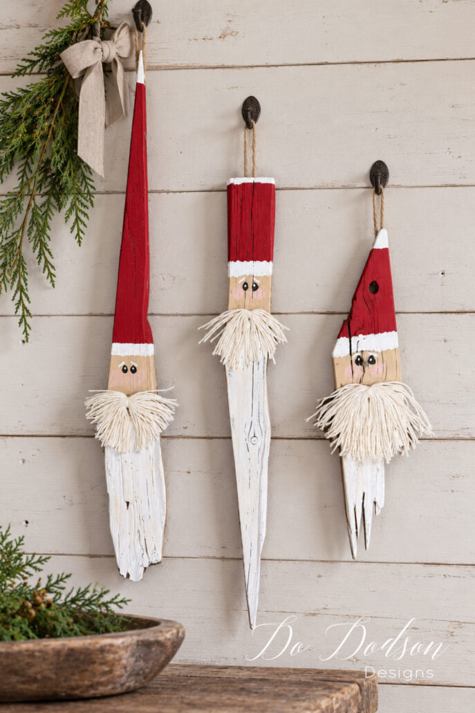 Three rustic scrap wood Santas hanging from jute loops on dark metal hooks against white shiplap, accented with cedar greenery and a linen bow.