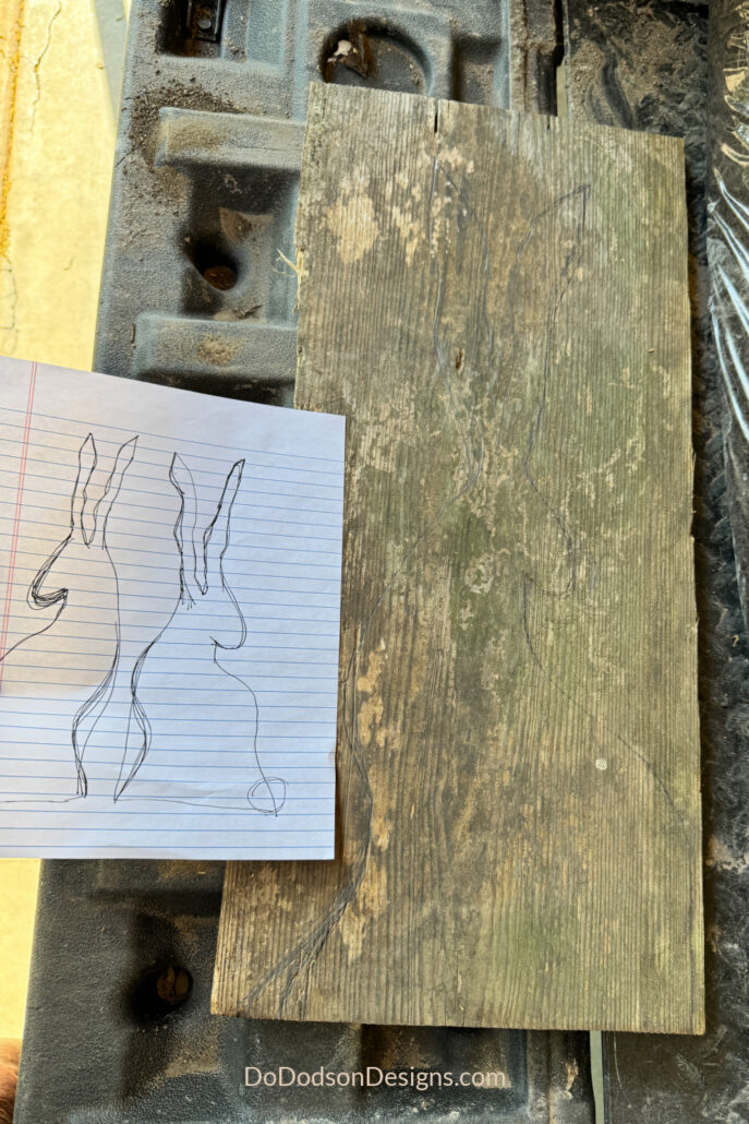 Scrap wood board with a hand-drawn Easter bunny outline traced onto the wood, next to a paper sketch of bunny silhouettes, ready for cutting in a DIY woodworking project.