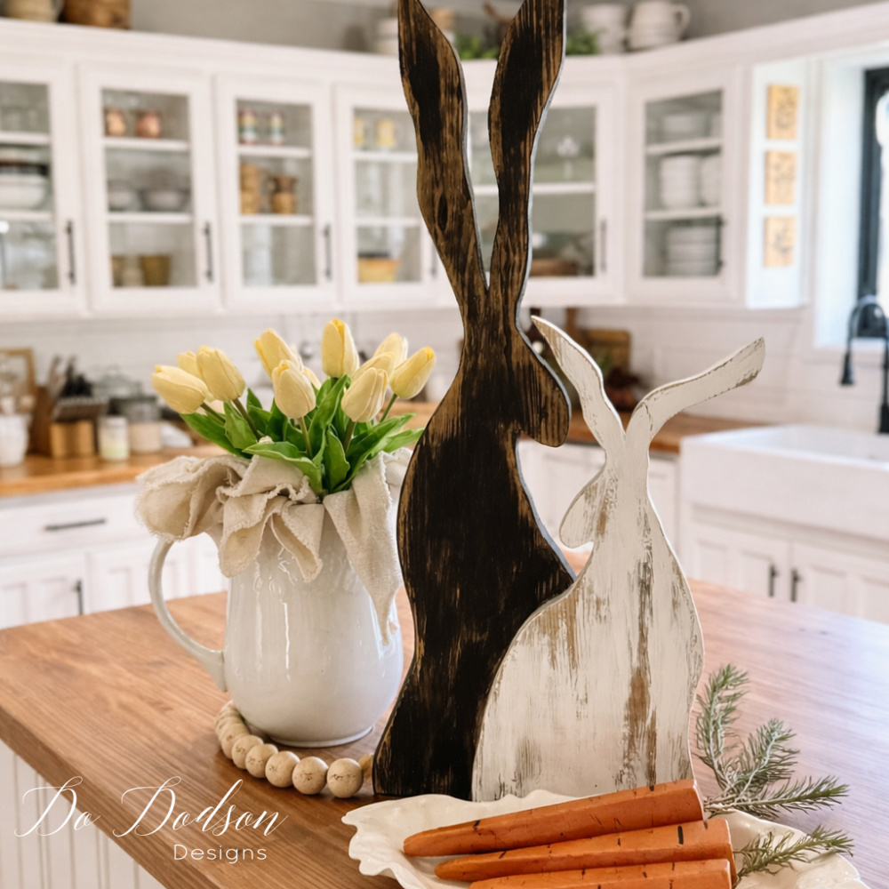 Rustic DIY wood Easter bunnies displayed on a farmhouse kitchen island with wooden carrots, tulips in a white pitcher, and neutral spring decor.