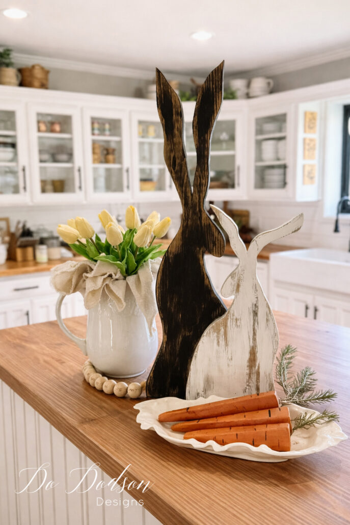 DIY wood Easter bunnies styled on a farmhouse butcher block island with painted wooden carrots, tulips in a white pitcher, and neutral spring decor in a bright kitchen.