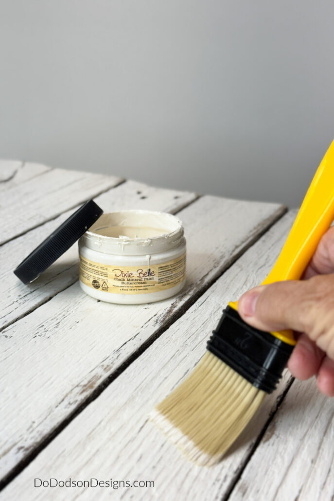 Applying Dixie Belle chalk mineral paint with a brush on a distressed white wood surface for a DIY wood sign project.