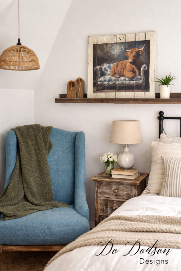 Farmhouse-style pallet wood wall art featuring a Highland cow design, displayed on a floating shelf with rustic decor and a blue chair below.