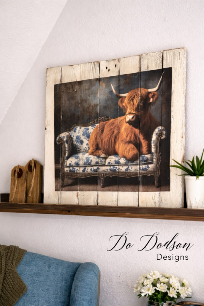 Rustic farmhouse wall art made from reclaimed pallet wood featuring a Highland cow on a vintage sofa, styled on a wooden shelf with simple decor.