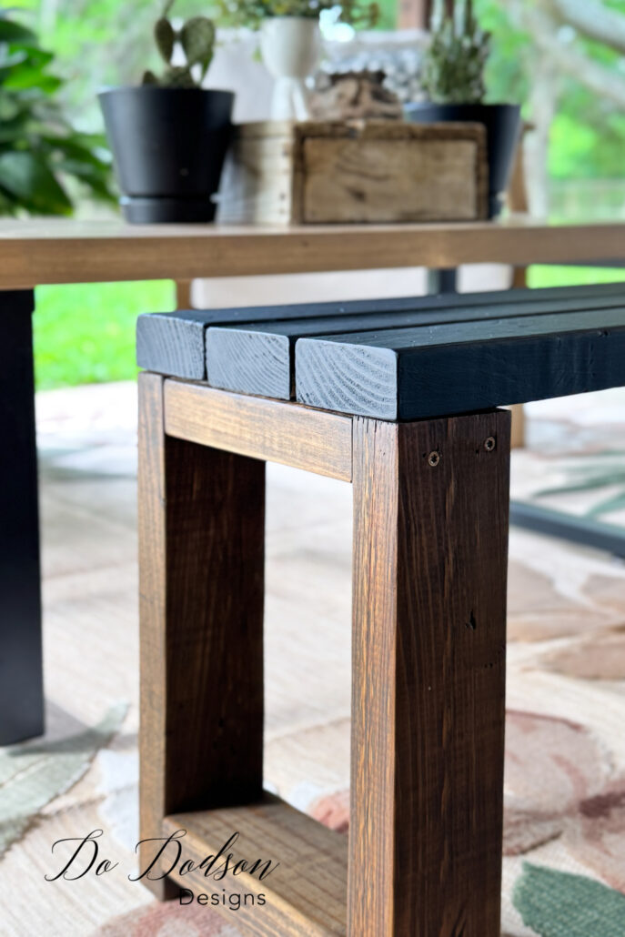 Rustic DIY wood bench made from 2x4 lumber with a dark stained base and black painted top, styled indoors near a table with potted plants, showing sturdy square legs and simple farmhouse design.