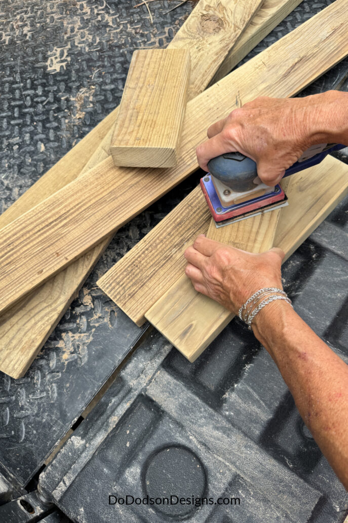 Sanding 2x4 wood boards with an orbital sander to smooth surfaces before assembling a DIY wood bench.
