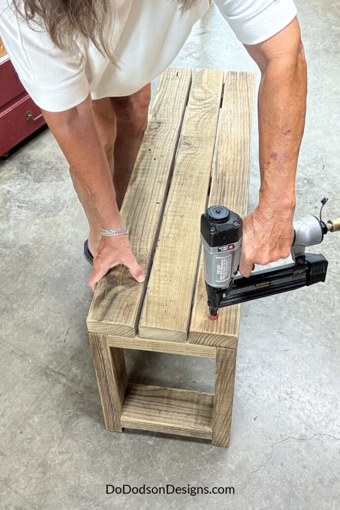 Attaching 2x4 boards to the top of a DIY wood bench using a nail gun, showing the bench seat being assembled.