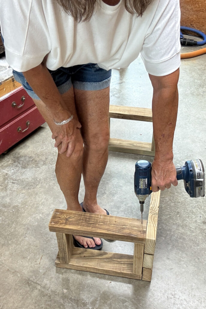 Drilling screws into a 2x4 frame to assemble the base of a DIY wood bench, showing the side frame being secured.