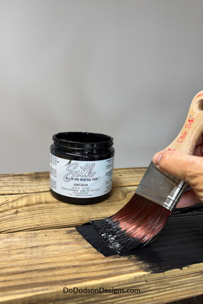 Applying black mineral paint to a 2x4 wood bench top using a paintbrush.