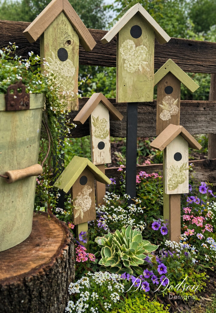 Close-up of DIY decorative birdhouse stakes in a flower bed with colorful blooms and rustic garden decor in a backyard setting.