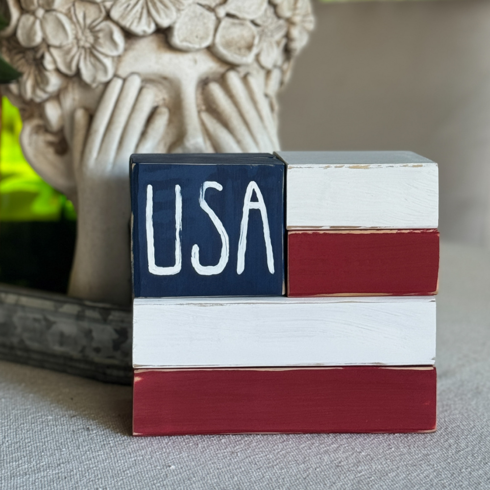 Handmade patriotic wood block flag displayed on a tabletop beside a decorative floral planter, featuring painted red, white, and blue scrap wood blocks for simple farmhouse-style 4th of July decor.
