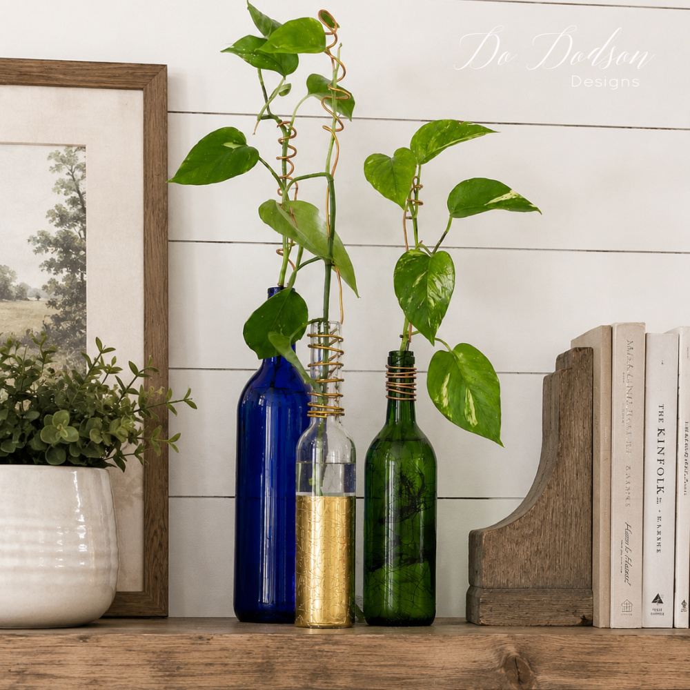 Pothos plant cuttings displayed in blue, green, and gold glass bottles on a rustic wooden shelf against a white shiplap wall with farmhouse decor accents including books, greenery, and framed artwork.