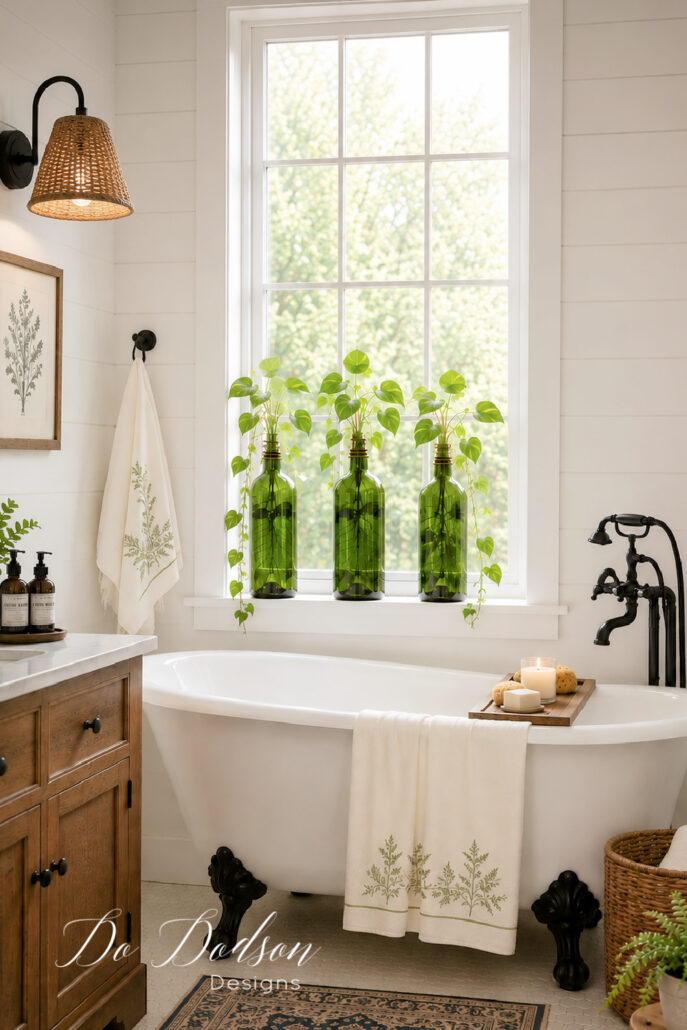 Bright modern farmhouse bathroom with a white clawfoot tub beneath a tall window featuring three green glass bottles filled with pothos plant cuttings, cream towels with green floral accents, a wicker shade wall sconce with black base, warm wood vanity, and soft natural light.