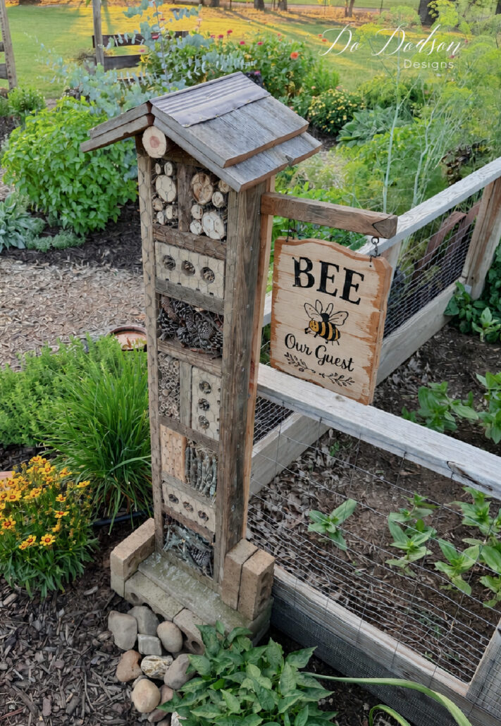 Rustic DIY bug house filled with natural nesting materials including wood slices, bamboo tubes, bark, and pinecones, standing beside a raised garden bed in a lush cottage garden. A handmade wooden sign reading &ldquo;Bee Our Guest&rdquo; hangs from the side, surrounded by colorful flowers, herbs, and pollinator-friendly plants, creating a welcoming habitat for bees and beneficial insects.