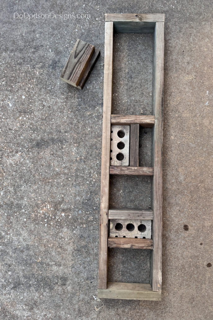 Early stages of a DIY bug house build showing a weathered wooden frame with divided compartments and concrete blocks on a patio. The insect hotel structure is being assembled with multiple sections designed to hold natural nesting materials for bees, butterflies, and beneficial garden insects.