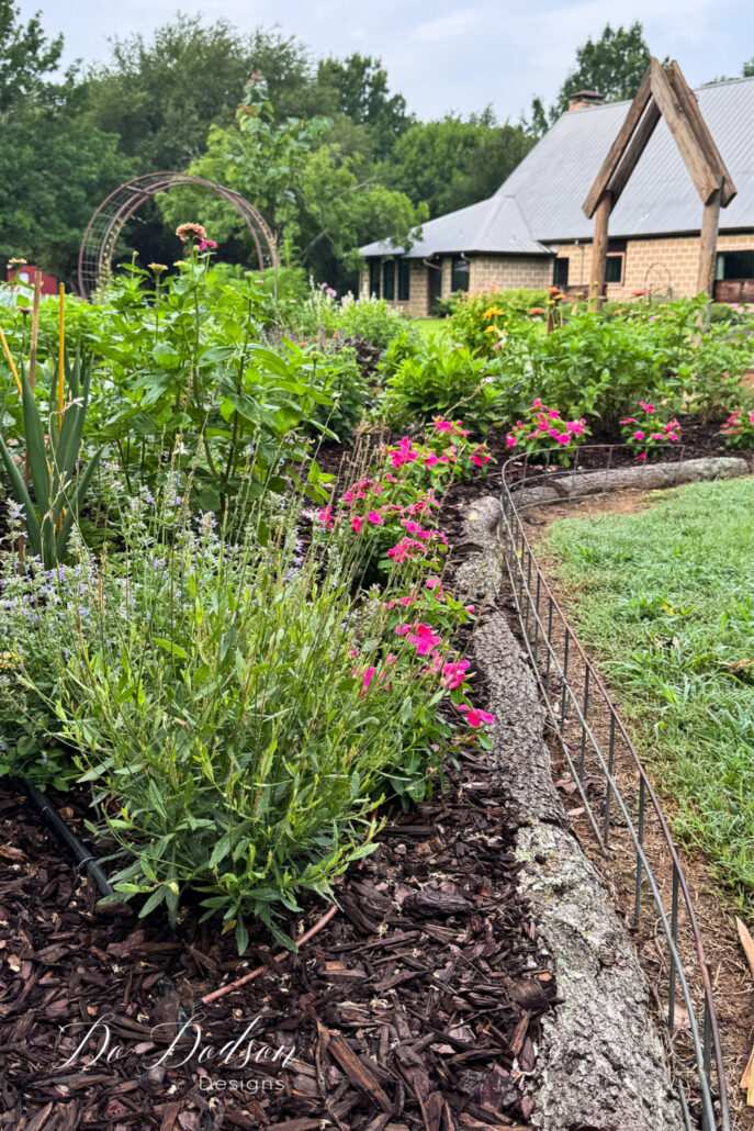 Colorful country cottage garden featuring curved flower beds filled with zinnias, lavender, and mixed perennials. A rustic arbor and wooden garden structure frame the backyard design, while layered plantings create depth and seasonal interest around a lush lawn and farmhouse setting.