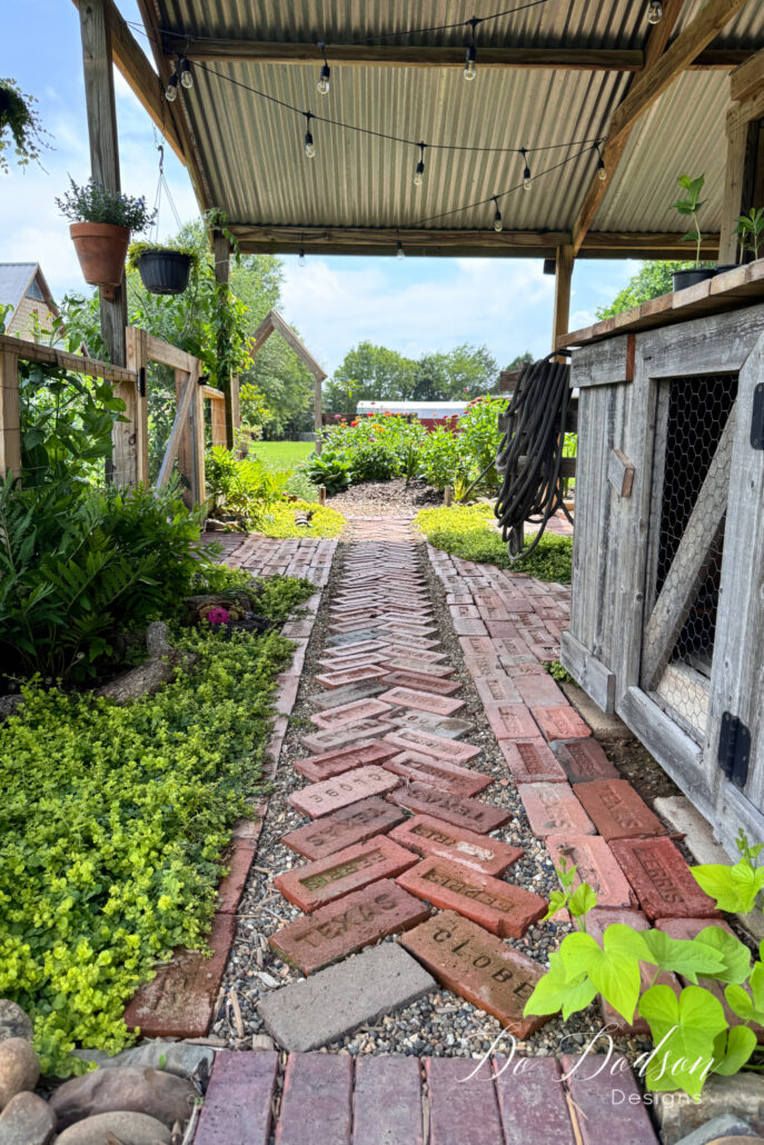 Antique brick garden pathway beneath a covered garden shed, bordered by lush chartreuse ground cover, ferns, and cottage garden plants. Reclaimed bricks laid in a herringbone pattern lead through the garden toward a sunny flower bed and wooden arbor in the distance. Hanging planters, string lights, a weathered potting cabinet, and climbing vines create a charming farmhouse garden retreat.