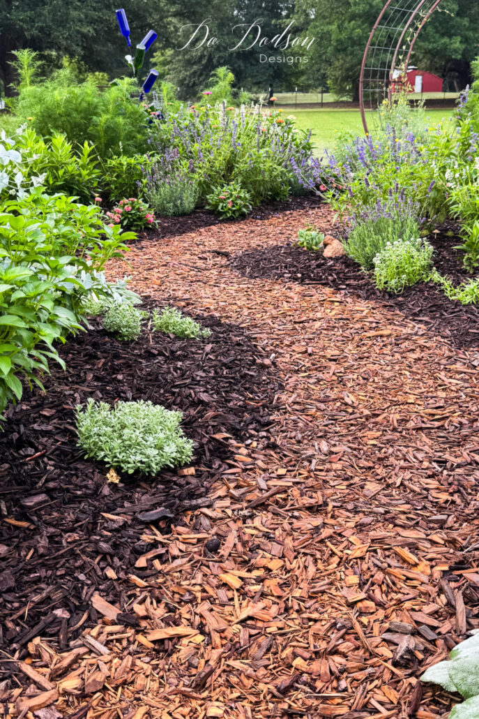 Curved mulch garden path winding through a colorful country cottage garden filled with lavender, flowering perennials, and lush greenery. A rustic metal arbor and blue bottle tree create charming focal points in this backyard garden design, surrounded by layered plantings and cottage-style landscaping.
