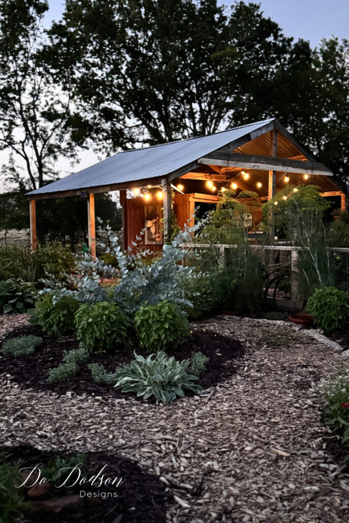Rustic country cottage garden at dusk featuring a winding mulch pathway bordered by lamb&rsquo;s ear, herbs, and cottage garden plants. A charming open-air garden shed glows with warm string lights, creating a cozy evening atmosphere among lush greenery, eucalyptus, and farmhouse-inspired garden beds.