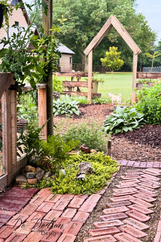 Rustic DIY wooden arbor positioned at the entrance to a cottage garden, framed by winding brick pathways, lush greenery, hostas, climbing vines, and cottage-style flower beds. The handcrafted arbor creates a charming focal point and inviting garden entrance.