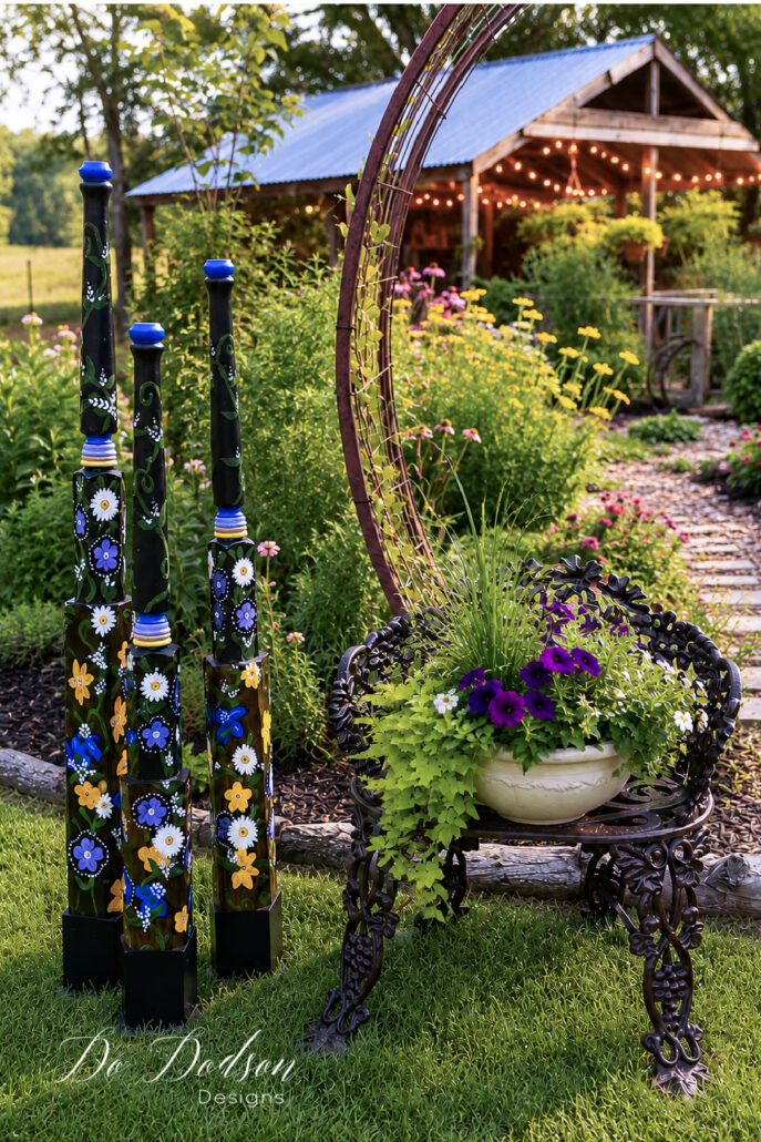 Hand-painted DIY garden totem poles stand beside an ornate metal garden bench holding a planter filled with lush summer greenery and purple flowers. Set in a vibrant cottage garden, the colorful floral-painted poles create a striking focal point beneath a rustic garden arch, while a charming garden shed glows with warm caf&eacute; string lights in the background.