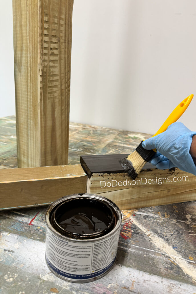 A gloved hand applies dark wood stain to reclaimed wood pieces for a DIY garden totem pole project. The recycled wood components are being refinished and prepared for assembly into hand-painted garden poles and homemade garden art made from scrap wood and thrifted materials.