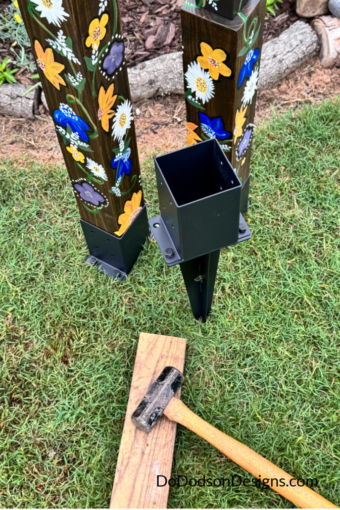 Ground anchors are being installed to secure hand-painted DIY garden totem poles in the landscape. A metal post anchor, hammer, and wood block are shown beside colorful floral-painted wood poles, demonstrating how to safely mount homemade garden art made from recycled materials, scrap wood, and thrifted finds for outdoor display.