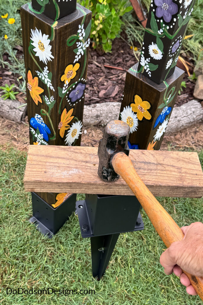A hammer and wood block are used to drive a metal ground anchor into the soil for installing DIY garden totem poles. The hand-painted wooden poles feature colorful floral designs and are secured with black post anchors, creating durable homemade garden art made from recycled materials, scrap wood, and thrifted finds for a cottage garden display.