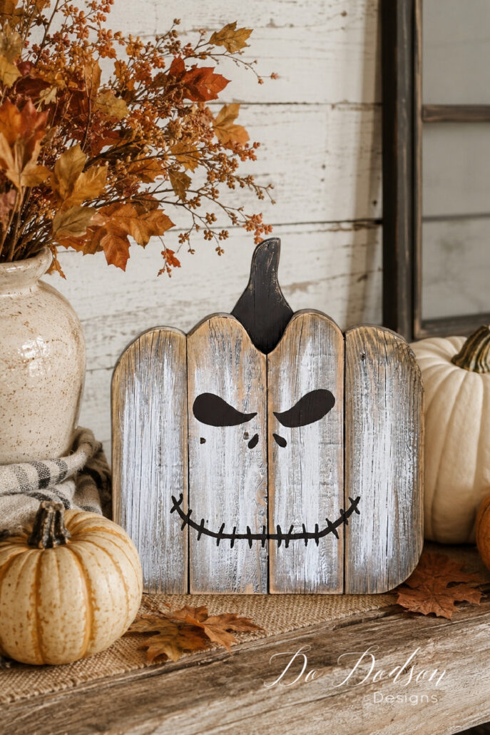 Rustic DIY wooden jack-o'-lantern decoration displayed on a weathered farmhouse table with white and orange pumpkins, autumn leaves, and a crock filled with fall branches against a white shiplap wall. The cozy neutral fall vignette creates a warm farmhouse Halloween display.