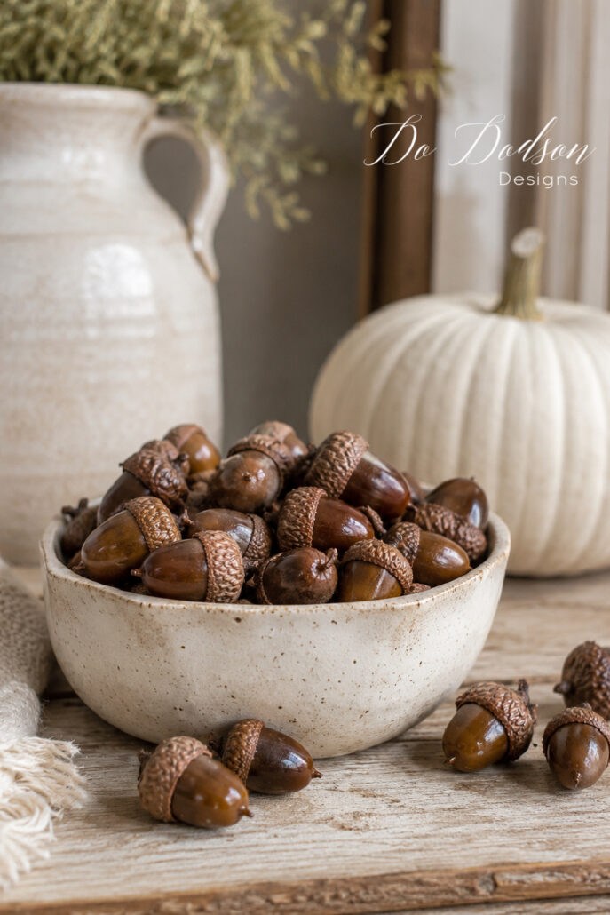 DIY preserved acorns displayed in a neutral stoneware bowl on a rustic farmhouse table. The glossy brown acorns are the focal point of the cozy fall vignette, styled with a cream ceramic crock, white heirloom pumpkin, soft linen fabric, and weathered wood for simple, neutral autumn farmhouse decor.