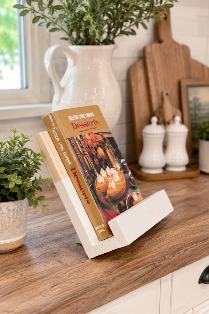DIY wooden cookbook stand displayed on a modern farmhouse kitchen countertop holding a vintage cookbook. The handmade stand is styled with a white ceramic pitcher, potted greenery, wooden cutting boards, and neutral kitchen decor, creating a warm and functional cooking space.