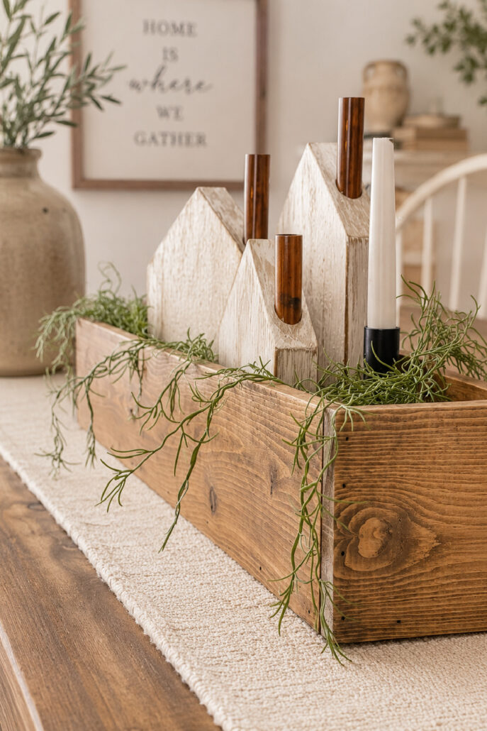 DIY wooden centerpiece box styled on a modern farmhouse dining table with white decorative wooden houses, copper pipe candle holders, pillar candles, and trailing greenery. The rustic wood box sits on a neutral woven table runner with soft natural light, creating a cozy farmhouse centerpiece perfect for year-round home decor.
