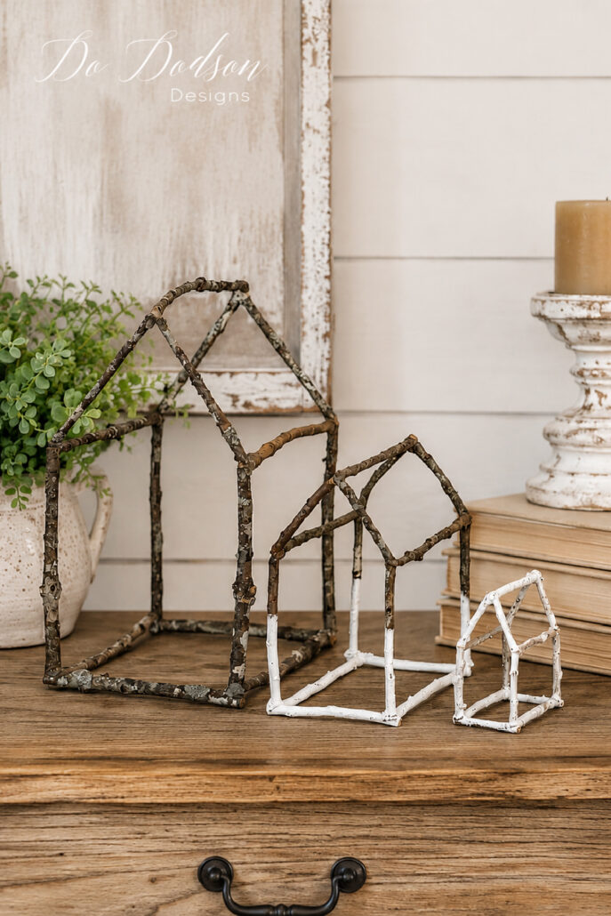 Three DIY twig houses made from natural branches are displayed on a rustic farmhouse table in a neutral home decor vignette. The handcrafted twig house sculptures are styled with a distressed picture frame, stacked vintage books, a weathered candlestick, and soft greenery, creating a cozy cottage-inspired farmhouse display.