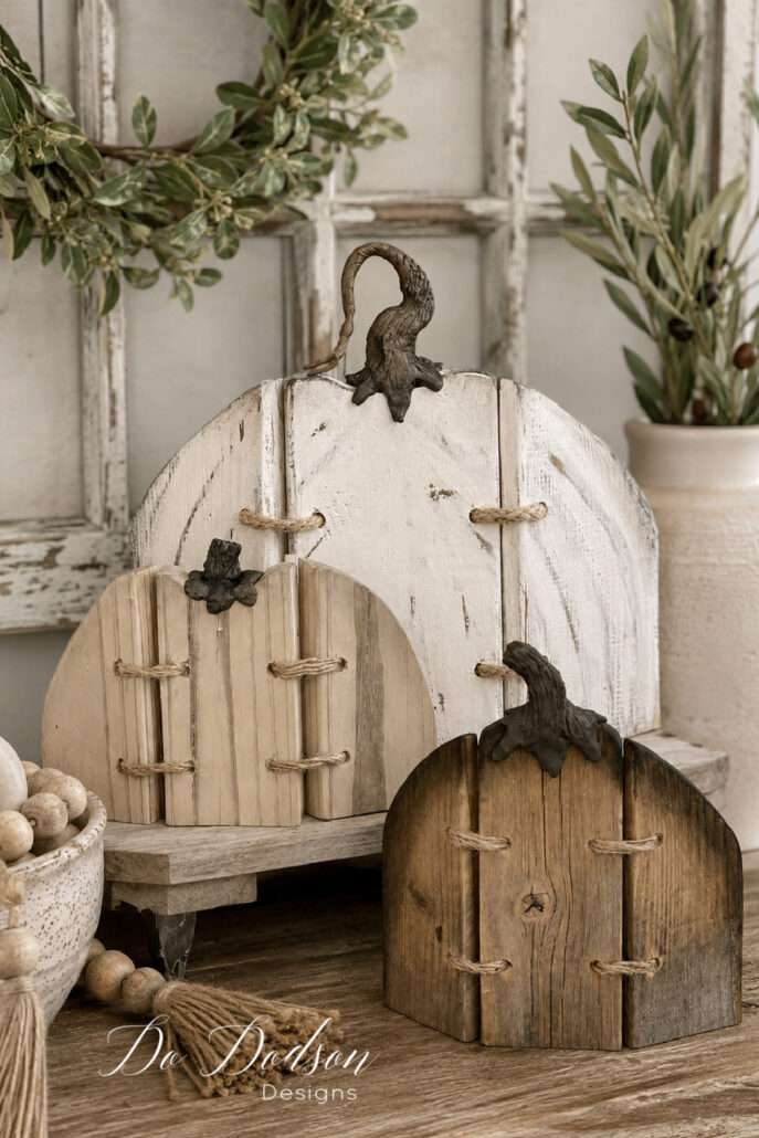 Three handcrafted rustic wood pumpkins displayed in a cozy neutral farmhouse fall vignette. The DIY wooden pumpkins feature weathered finishes, natural wood grain, twine lacing, and realistic pumpkin stems, styled with a distressed vintage window frame, olive greenery, a ceramic vase, wooden bead garland, and warm wood accents for simple autumn home decor.