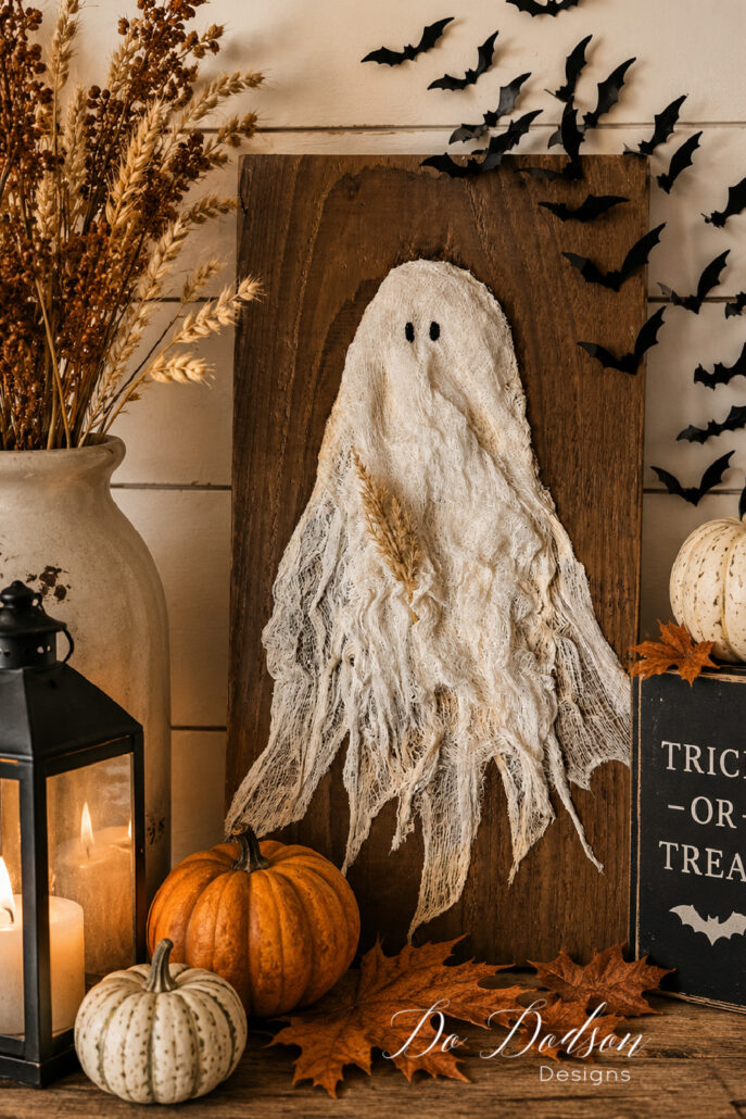 DIY cheesecloth ghost wall hanging displayed in a cozy farmhouse Halloween vignette with rustic wood, vintage lanterns, white and orange pumpkins, dried wheat, black bat decorations, and colorful fall leaves. The handmade ghost is mounted on a weathered wooden board, creating charming neutral Halloween decor.