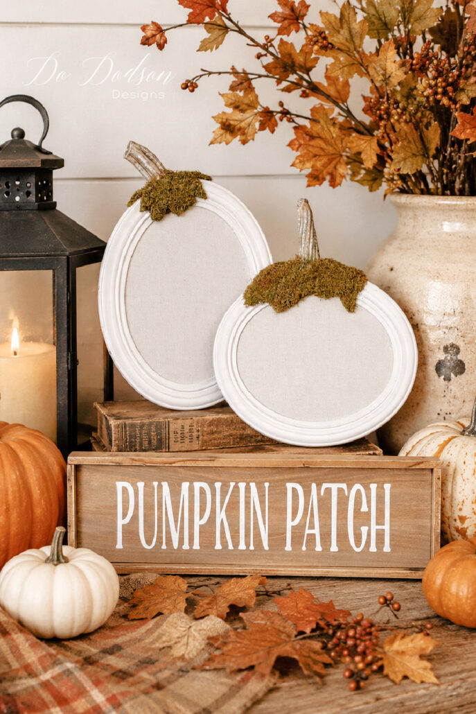 DIY pumpkin picture frames displayed in a cozy farmhouse fall vignette with a rustic Pumpkin Patch sign, vintage lantern, cream crock filled with autumn branches, stacked antique books, and white and orange pumpkins. Moss-covered pumpkin stems and colorful fall leaves complete the warm harvest display.