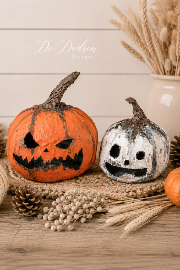Two handmade DIY paper mache pumpkin heads with painted jack-o'-lantern faces are displayed on a rustic wooden table in a cozy farmhouse setting. The orange pumpkin features a spooky carved-style grin, while the white pumpkin has a playful ghost-like face. Pinecones, dried wheat, neutral berries, and a cream stoneware vase with dried grasses create a simple, rustic fall display against a white shiplap wall.