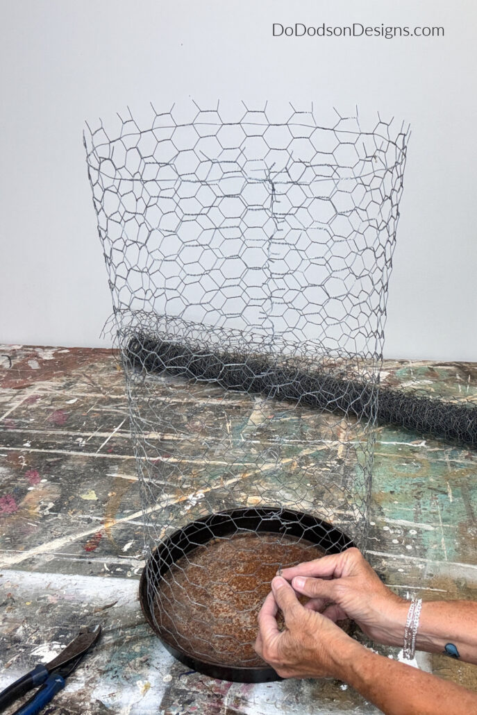 Hands joining the seam of a DIY chicken wire cloche over a vintage metal tray during assembly. The step-by-step garden project uses a 38-by-28-inch piece of chicken wire to create a rustic protective cage for flowers, herbs, and vegetable seedlings.