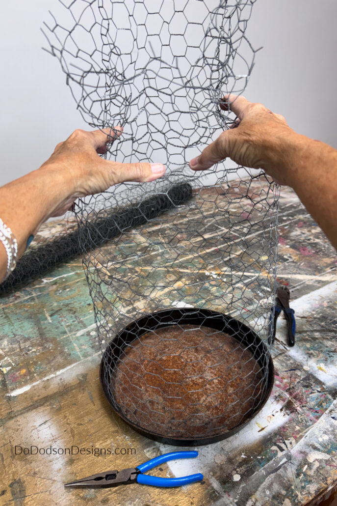 Hands gently gathering the top of a DIY chicken wire cloche to form a rounded dome over a vintage metal tray. This step in the garden cloche DIY project shapes the wire cage before twisting the top closed to create a decorative plant protector for flowers, herbs, and vegetables.