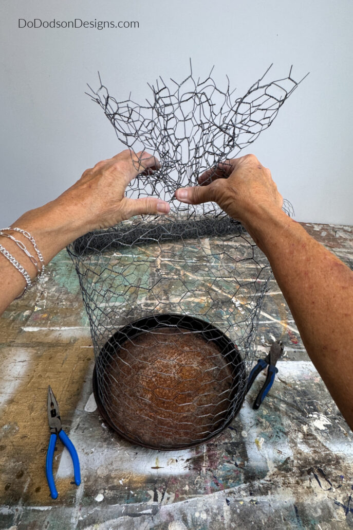 Hands gathering and twisting the top of a DIY chicken wire cloche to create its decorative knob. This step in the garden cloche DIY project transforms a simple chicken wire cage into a charming plant protector for flowers, herbs, vegetables, and cottage-style garden décor.