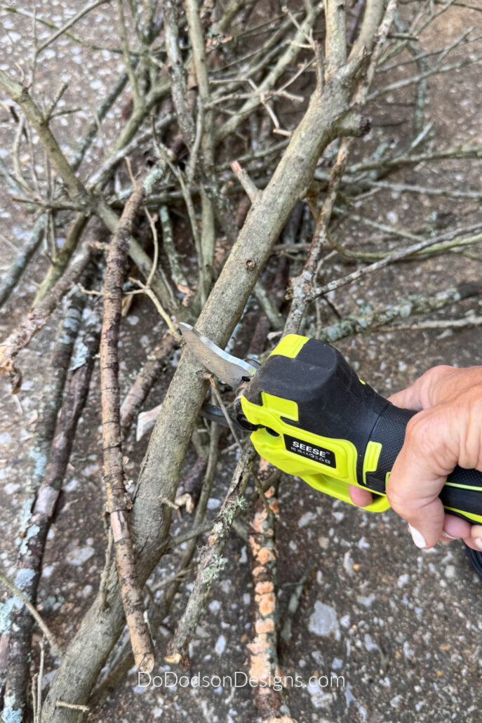 Trimming tree branches with battery operated pruning shears to make a rustic DIY witch&rsquo;s broom from natural twigs.