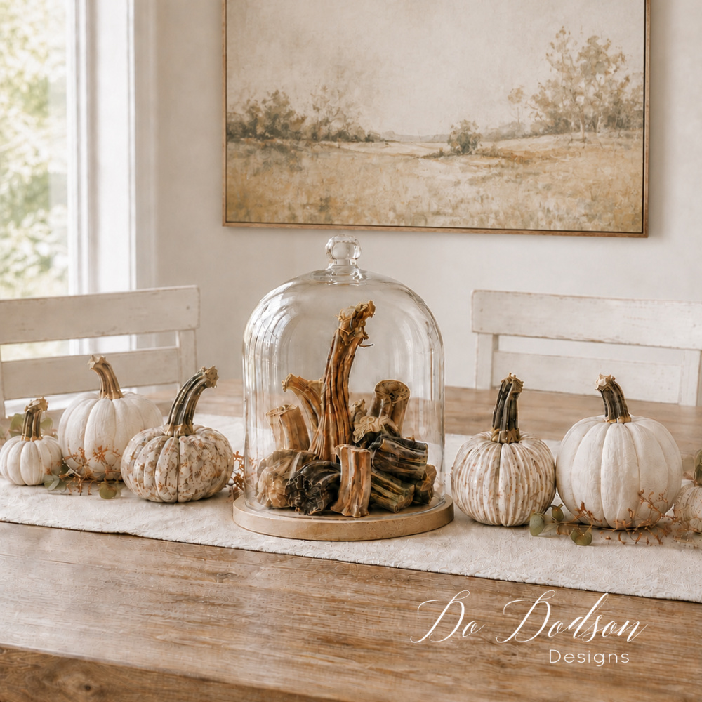 Real dried pumpkin stems displayed under a glass cloche with white and floral fabric pumpkins on a neutral fall dining table.