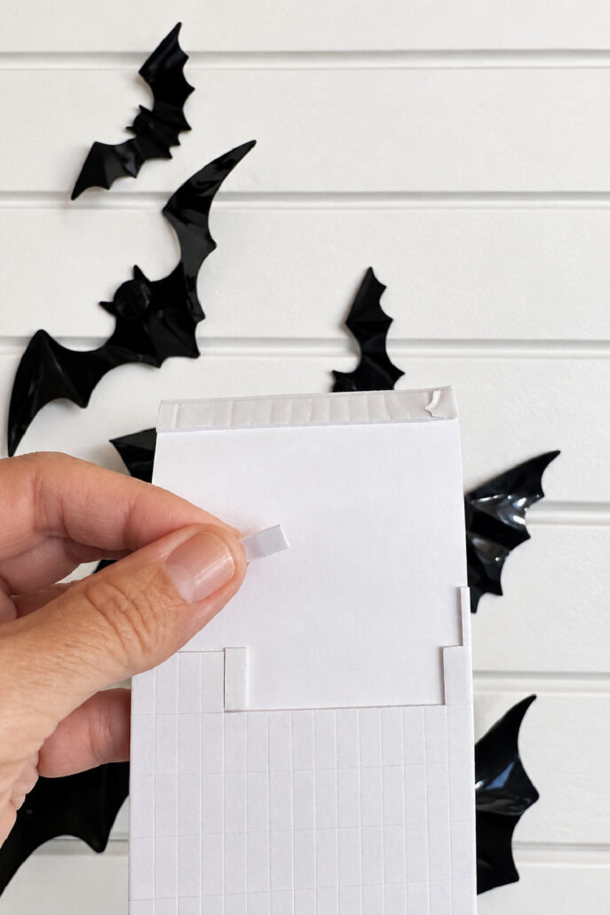 Hand holding double-sided adhesive tape used to attach 3D PVC bats to a Halloween bat wall.